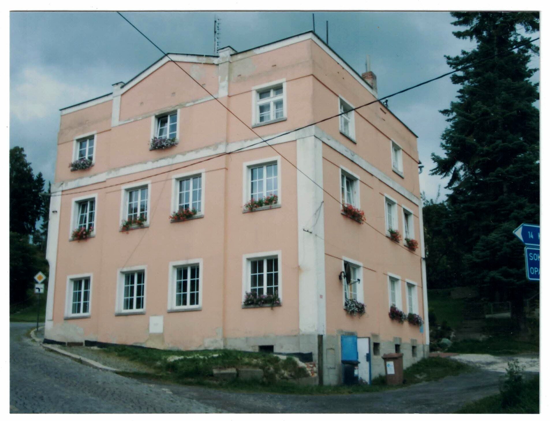 白い窓枠と花箱のある3階建ての桃色がかった角地の住宅のカラー写真。曇り空の下の石畳通り、Sokolov 方向を示す青い道路標識。