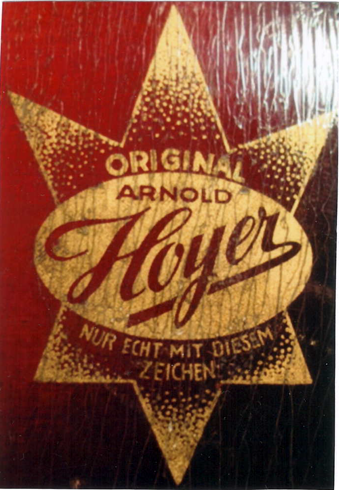 赤みがかった木目に金押しされた真贋ラベルの接写。楕円内に「ORIGINAL ARNOLD Hoyer」と多角星、ドイツ語の一文「NUR ECHT MIT DIESEM ZEICHEN.」。